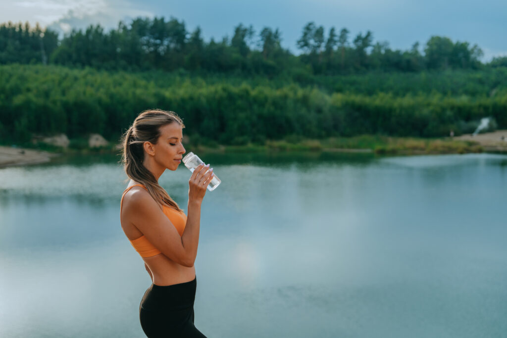 Peaceful sporty Swedish young woman in sportswear holds bottle of water stands against lake, forest